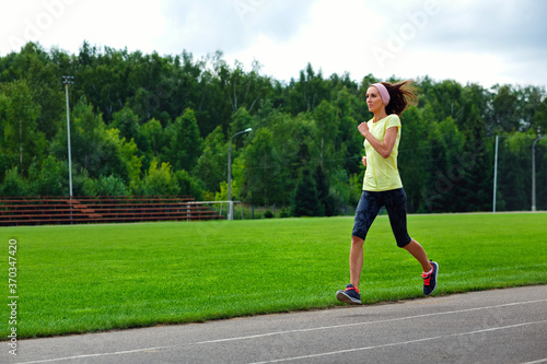 A beautiful fitness girl in a yellow sports top and leggings is Jogging around the stadium. She runs fast on a warm summer day. The Sportswoman Is Engaged In Her Usual Sports Practice.