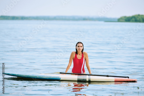 Happy young brunette woman in red swimsuit standing in water by surfboard
