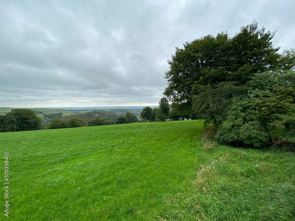 Landscape view, with heavy clouds, over a green meadow, with trees and hills in the distance near, Halifax, Yorkshire, UK