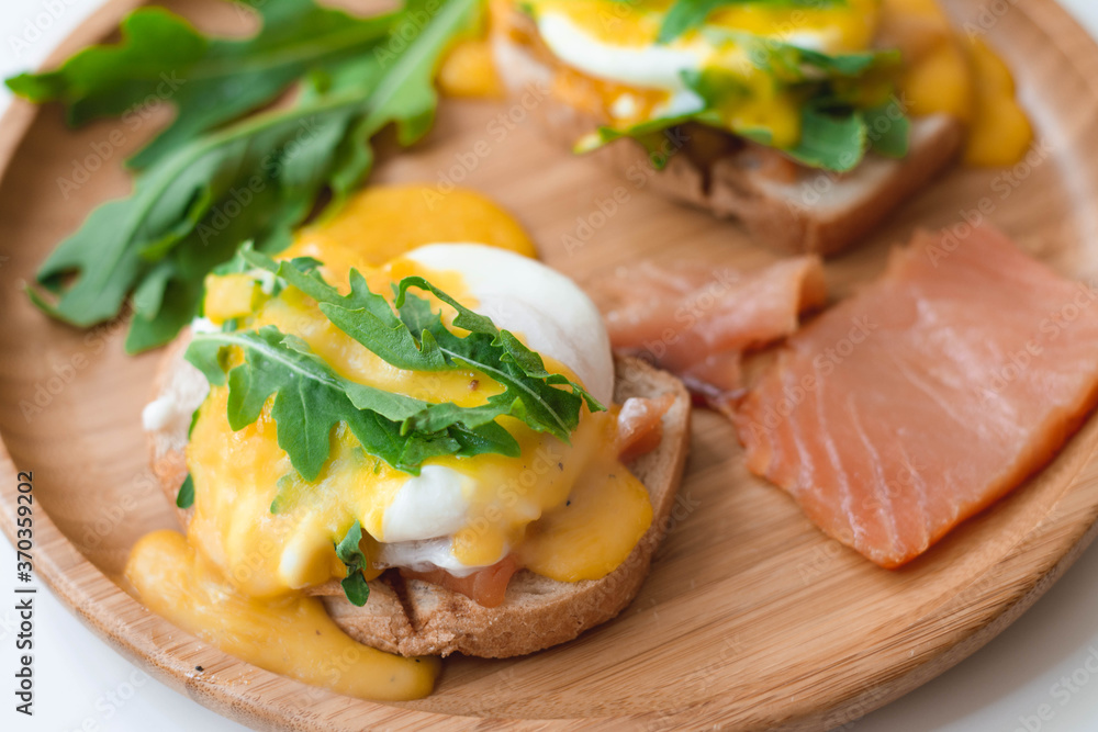 Poached eggs Benedict with Hollandaise sauce, smoked salmon, arugula on toasted bread. Wooden plate, white background.