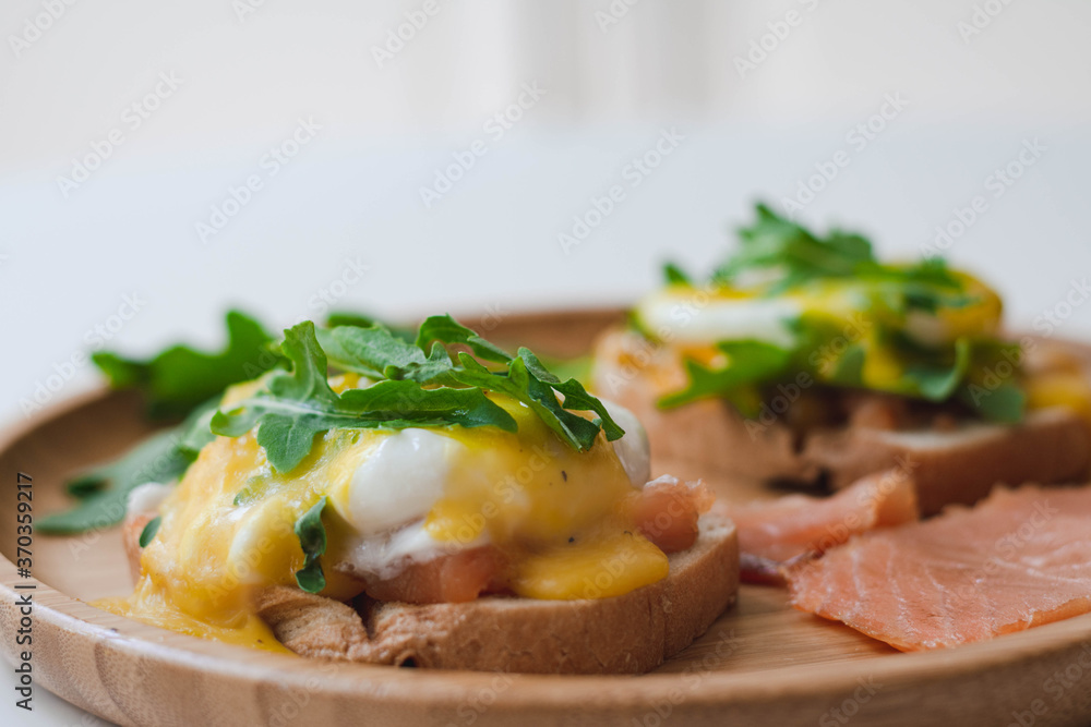 Poached eggs Benedict with Hollandaise sauce, smoked salmon, arugula on toasted bread. Wooden plate, white background. Poached eggs Benedict with Hollandaise sauce, smoked salmon, arugula on toasted bread. Wooden plate, white background.
