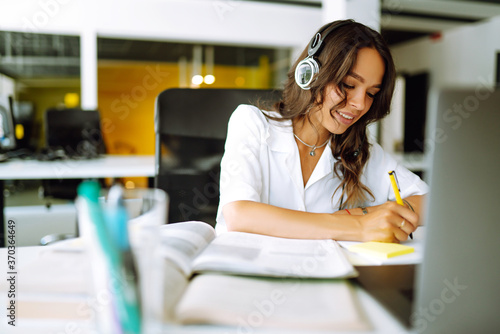 Young woman student wear wireless headphone study online. She learn language listen lecture watch webinar write notes look at laptop, distant education. Video call self-isolation during ncov situation photo