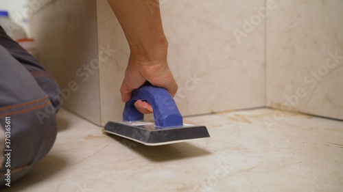 The hand of man holding a rubber float and filling joints with grout. The worker is rubbing the tiles with a float.