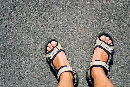 Tanned male legs in hiking sandals on the asphalt on a sunny day.