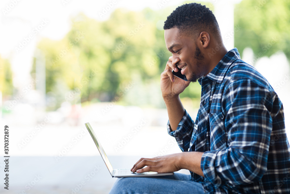 Black man using laptop, sitting in the city with cellphone Black man using laptop, sitting in the city with cellphone