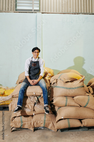 Happy young Asian man in apron sitting on sacks with roasted coffee
