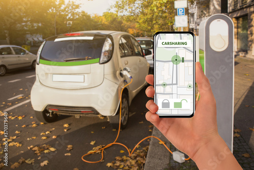 Cuadro en lienzo Hand with a mobile phone on the background of a charging carsharing electric car