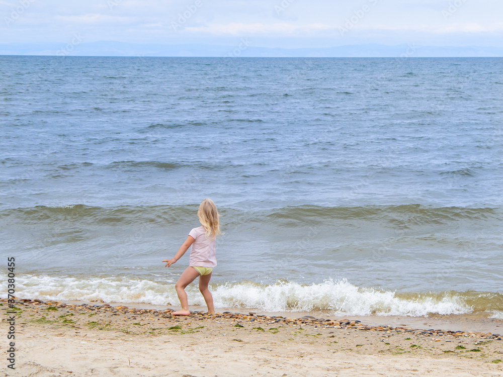 A little girl stands on the shore by the water, viewed from behind