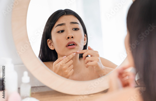 Upset asian girl checking for pimples  looking at mirror