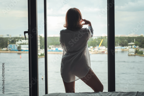 Back view of beautiful young woman in a transparent nightgown standing and enjoying near open panoramic window of apartments.