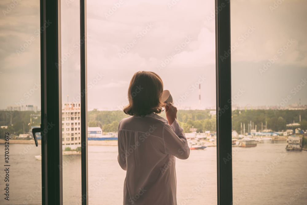 Back view of beautiful young woman with cup of coffee in a transparent nightgown standing and enjoying near open panoramic window of apartments. Back view of beautiful young woman with cup of coffee in a transparent nightgown standing and enjoying near open panoramic window of apartments.
