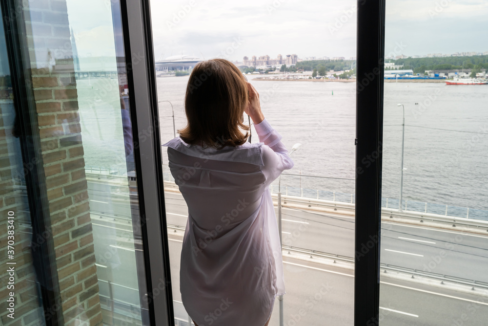 Back view of beautiful young happy woman in a transparent nightgown standing and enjoying near open panoramic window of skyscraper apartments.