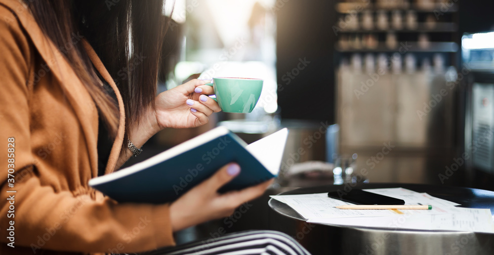 Young asian woman holding coffee cup to drink and read marketing plan in book while she work and relax in cafe, Selective focus of hand holding coffee cup