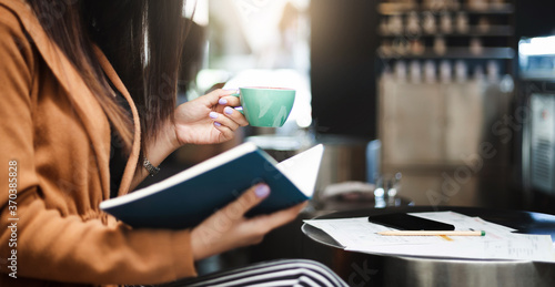 Young asian woman holding coffee cup to drink and read marketing plan in book while she work and relax in cafe, Selective focus of hand holding coffee cup