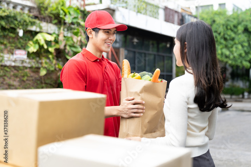 Asian delivery man wearing red uniform, red hat holding paper bag of food, fruit, vegetable give to female customer near parcel boxes. Concept of express delivery, food delivery, quarantined