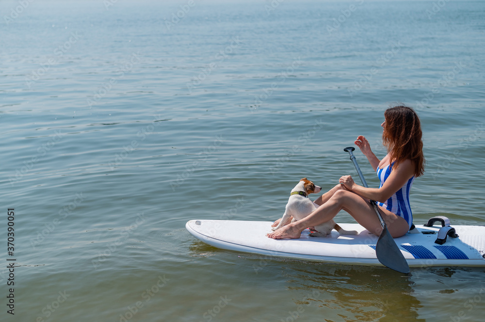 Fototapeta premium A woman is riding a sup surfboard with a dog on the lake. The girl goes in for water sports with her pet.