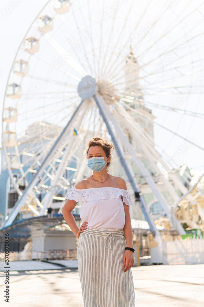 A beautiful young woman in a mask on the street is posing on a square near a ferris wheel and looking seriously on a sunny summer day