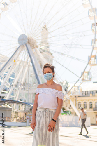 A beautiful young woman in a mask on the street is posing on a square near a ferris wheel and looking seriously on a sunny summer day