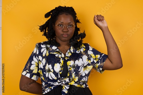 Foto Young pretty woman feeling serious, strong and rebellious, raising fist up, protesting or fighting for revolution against yellow wall