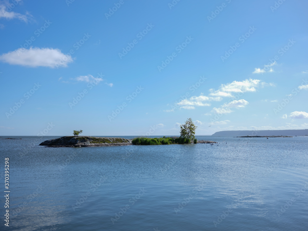 View of a rocky island with two isolated trees in a lake in Sweden. It is a clean image with calm waters and a blue sky background with a few clouds. The photograph is shot with a medium format camera