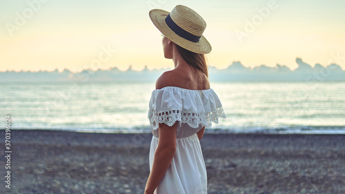 Romantic woman in a summer white dress with bare shoulders and a straw hat standing back by the seashore and enjoying serene and calm at summertime