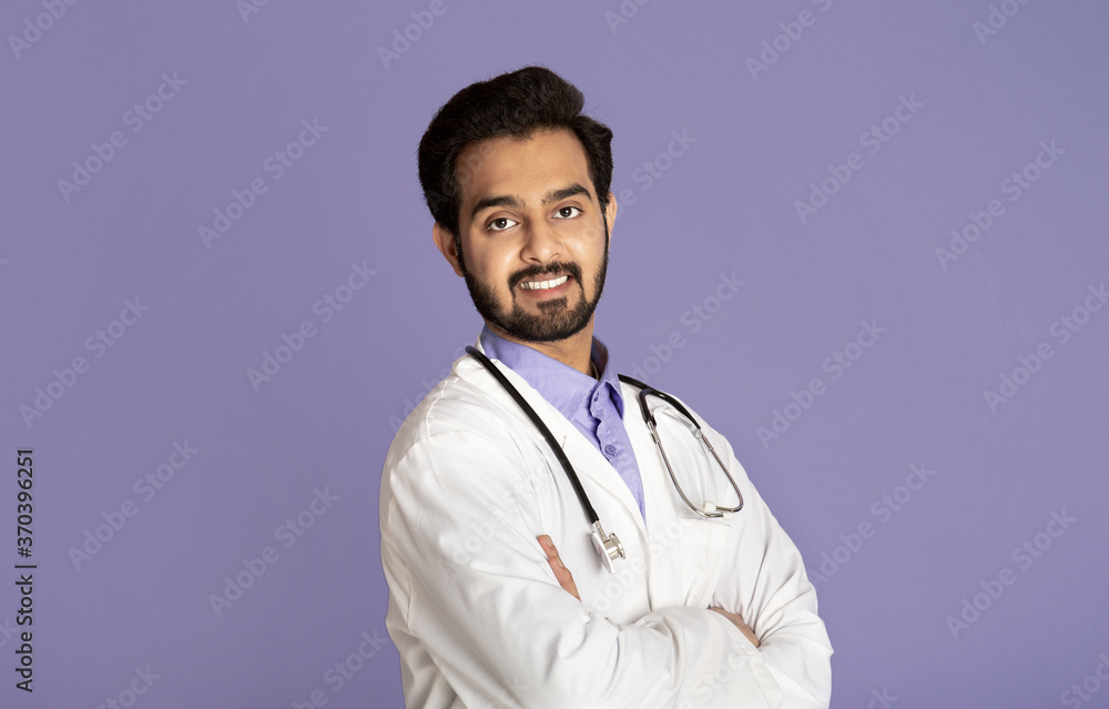 Smiling Indian doctor in lab coat standing with crossed arms on violet background Smiling Indian doctor in lab coat standing with crossed arms on violet background