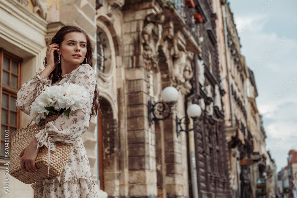Young elegant woman wearing floral print dress, holding wicker bag with white peony flowers, posing in street of European city. Lifestyle, travel conception. Copy, empty space for text
