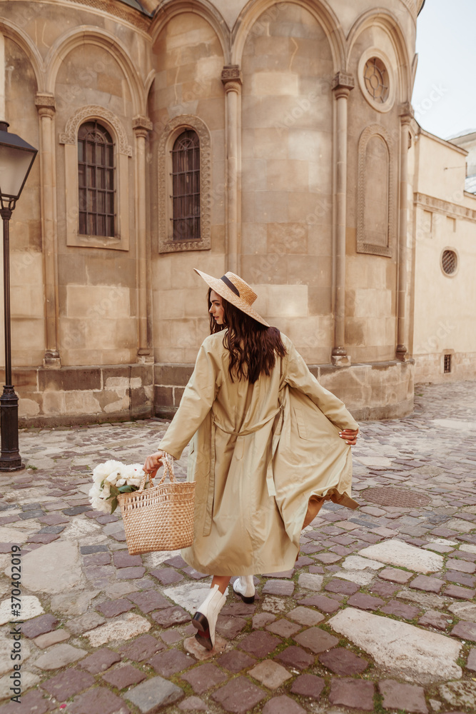 Lifestyle, travel conception: woman wearing straw hat, long trench coat, holding wicker bag with flowers, walking in street of European city