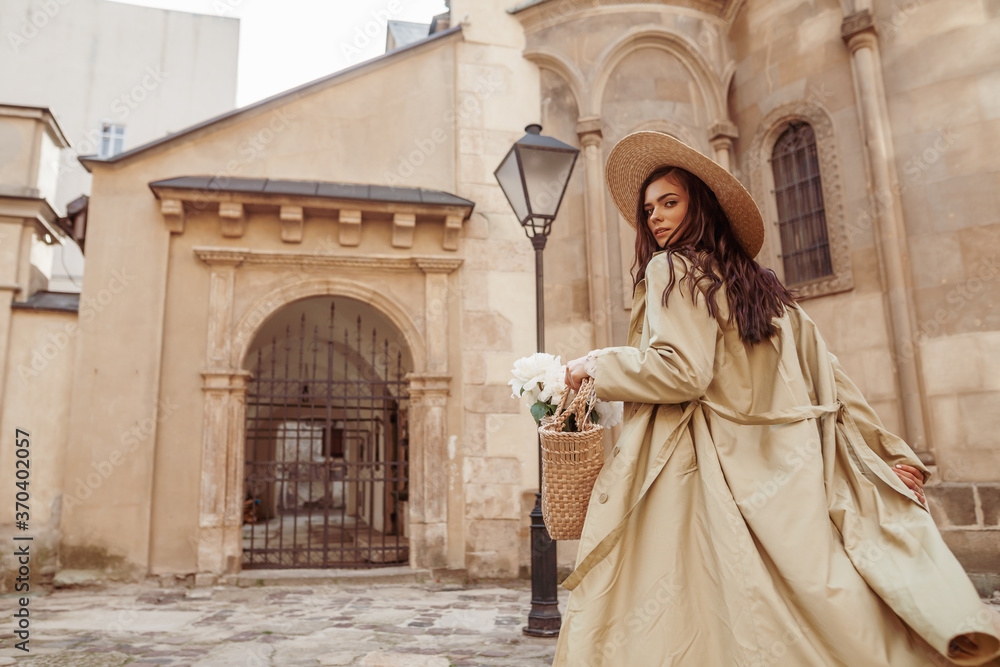 Lifestyle, travel conception: woman wearing straw hat, long trench coat, holding wicker bag with flowers, walking in street of European city. Copy, empty space for text