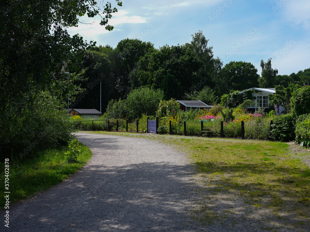 View of a dirt road with entrance to a garden area with cottages in Sweden. It is a hot summers day and the garden is in full bloom.