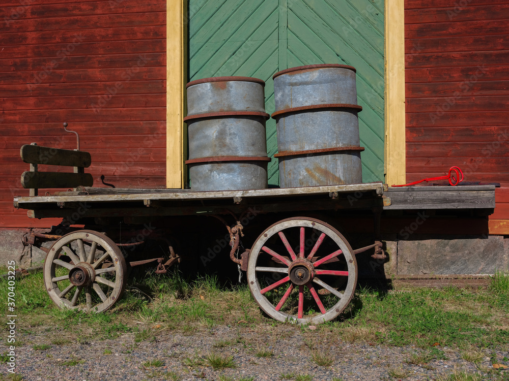 An old cart with two barrels standing in front of an old red barn with a green door. It is a sunny summers day.