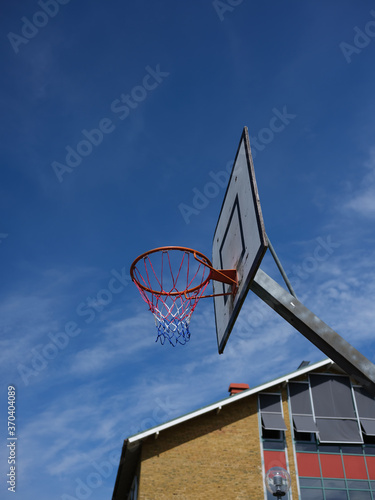 Basket hoop outside on a sunny day against a blue sky. Part of a building is shown in the background