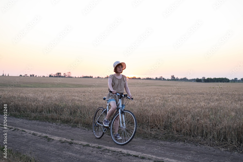Fototapeta premium Young woman with hat ride on the bicycle in summer wheat fields