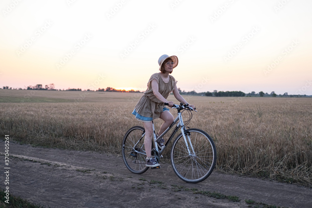 Fototapeta premium Young woman with hat ride on the bicycle in summer wheat fields