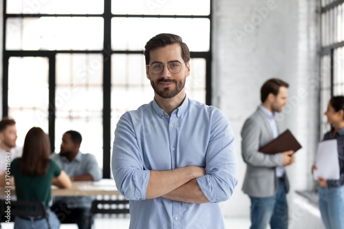 Head shot portrait smiling confident businessman wearing glasses standing in modern office room with arms crossed, diverse colleagues on background, executive boss startup founder looking at camera
