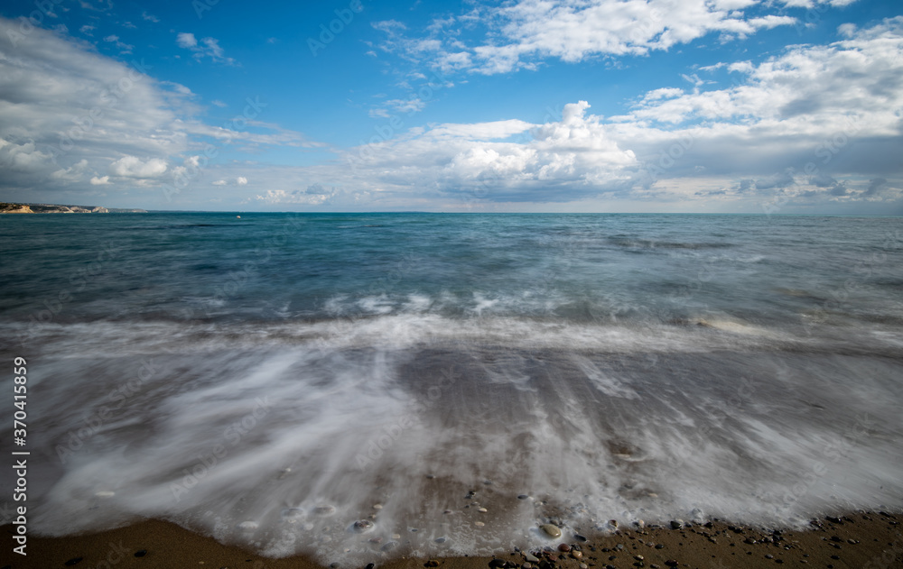 Seawaves splashing onto the sandy coast with pebbles and blue cloudy sky.