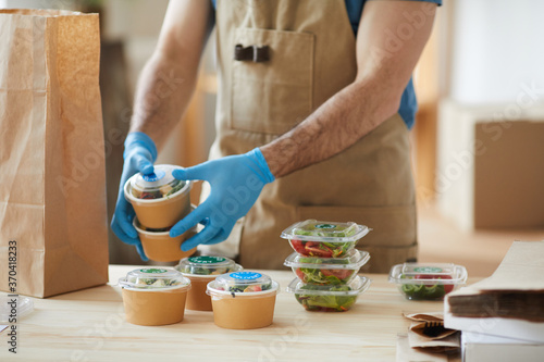 Closeup of unrecognizable worker wearing protective gloves packaging orders at wooden table in food delivery service, copy space