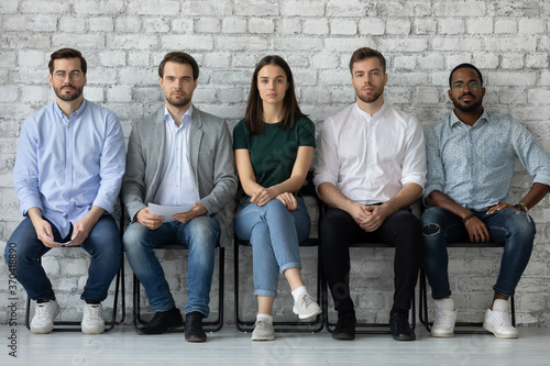Portrait of confident diverse candidates sitting in row in queue, serious applicants business people waiting for job interview, looking at camera, employments and recruitment concept