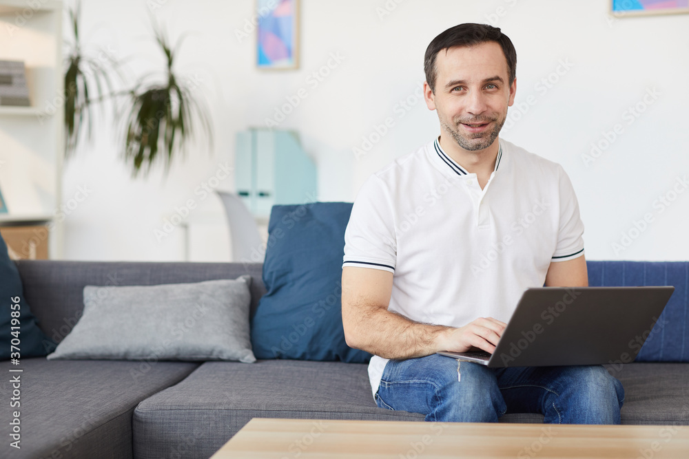 Portrait of smiling mature man using laptop and looking at camera while sitting on couch at home or in office, copy space
