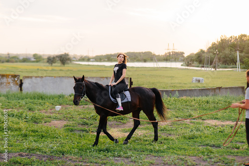 A young and pretty girl is learning to ride a thoroughbred Mare on a summer day at the ranch. Horse riding, training and rehabilitation