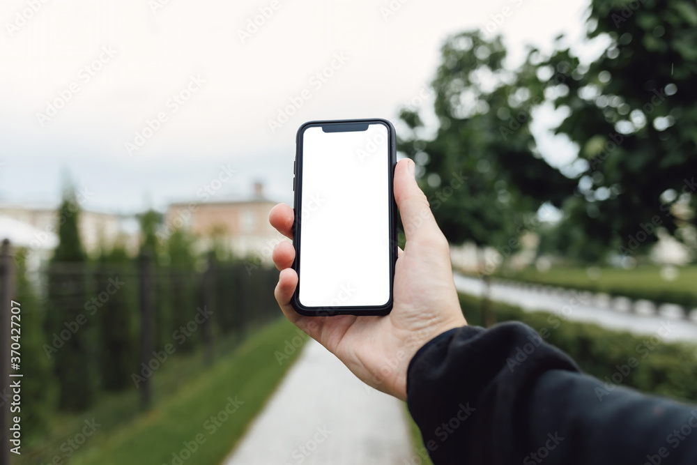 Mockup image of hand holding mobile phone with blank white screen. Man with a smartphone against the background of a city park with trees and a path.