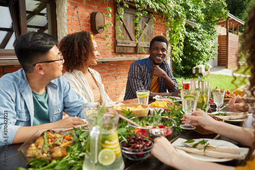 Portrait of multi-ethnic group of friends enjoying dinner together outdoors while sitting at table on open terrace, focus on smiling African-American man sharing stories, copy space