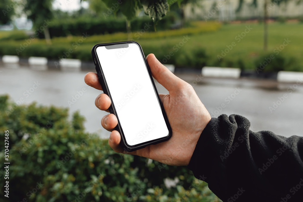 Mockup image of hand holding mobile phone with blank white screen. A man with a smartphone against the background of a city park with trees, green bushes and a road.