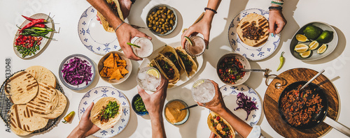 Friends having Mexican Taco dinner. Flat-lay of beef tacos, tomato salsa, tortillas, beer, snacks and peoples hands clinking glasses over white table, top view. Mexican cuisine, comfort food concept photo