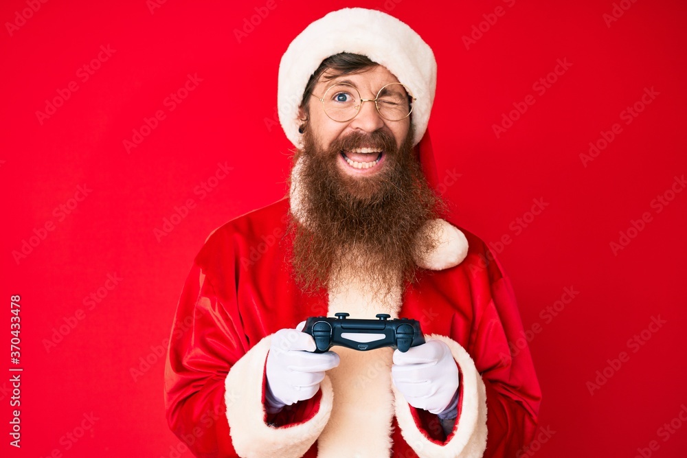 Handsome young red head man with long beard wearing santa claus costume playing video games winking looking at the camera with sexy expression, cheerful and happy face. Handsome young red head man with long beard wearing santa claus costume playing video games winking looking at the camera with sexy expression, cheerful and happy face.