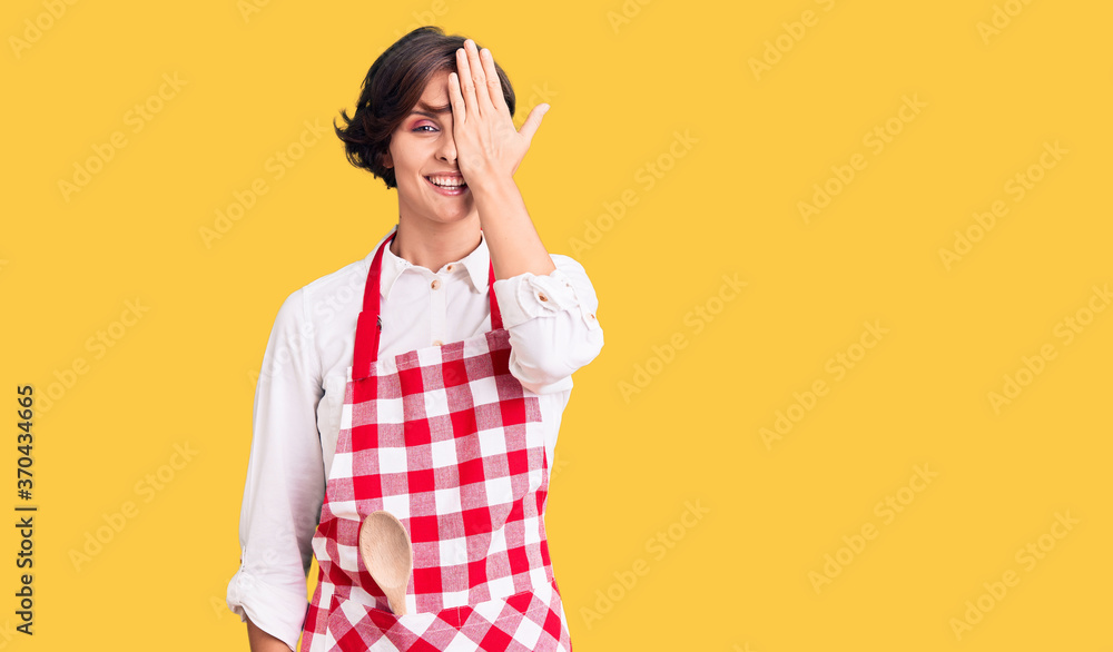 Beautiful young woman with short hair wearing professional cook apron covering one eye with hand, confident smile on face and surprise emotion. Beautiful young woman with short hair wearing professional cook apron covering one eye with hand, confident smile on face and surprise emotion.