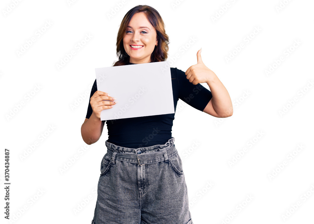 Young beautiful caucasian woman holding blank empty paper smiling happy and positive, thumb up doing excellent and approval sign Young beautiful caucasian woman holding blank empty paper smiling happy and positive, thumb up doing excellent and approval sign