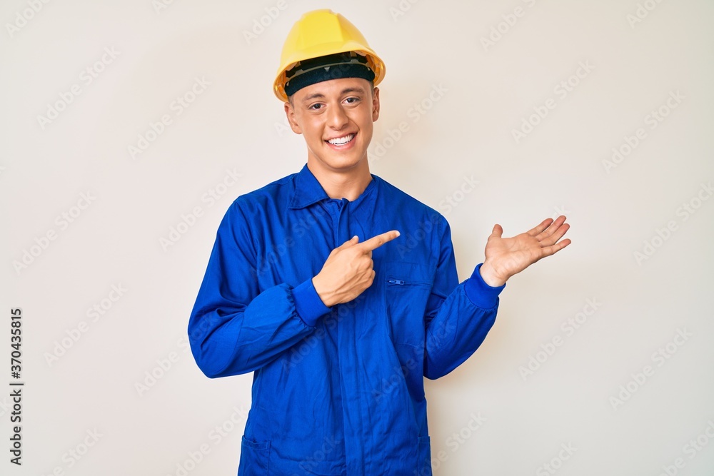 Young hispanic boy wearing worker uniform and hardhat amazed and smiling to the camera while presenting with hand and pointing with finger.