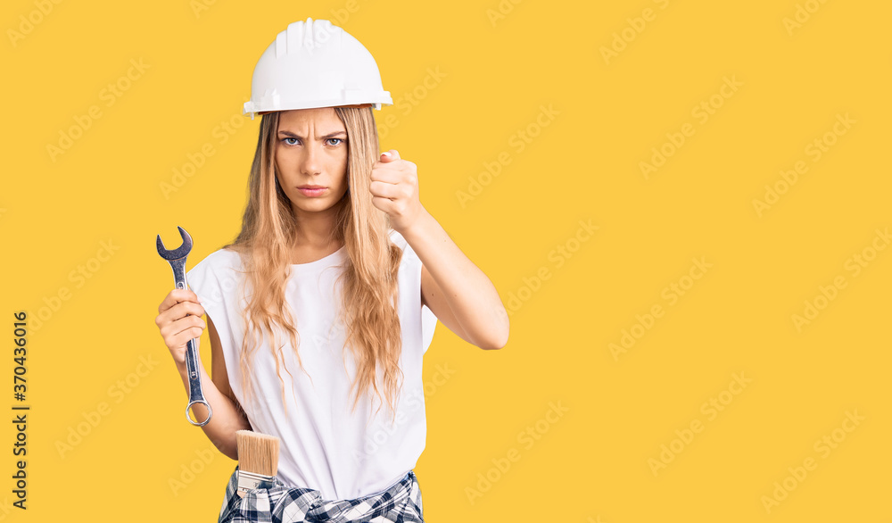 Beautiful caucasian woman with blonde hair wearing hardhat and builder clothes annoyed and frustrated shouting with anger, yelling crazy with anger and hand raised