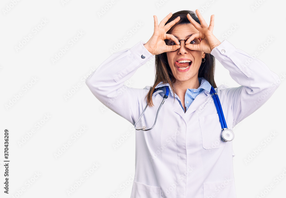 Young beautiful woman wearing doctor stethoscope and glasses doing ok gesture like binoculars sticking tongue out, eyes looking through fingers. crazy expression. Young beautiful woman wearing doctor stethoscope and glasses doing ok gesture like binoculars sticking tongue out, eyes looking through fingers. crazy expression.